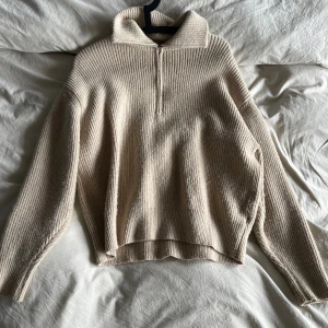 Beige stickad tröja med krage - Säljer en beige stickad tröja med ribbad struktur och hög krage. Tröjan har lång ärm och en relaxed passform. Perfekt för lager-på-lager och chill höst- eller vinterstil. Halv dragkedja framtill för extra detalj.