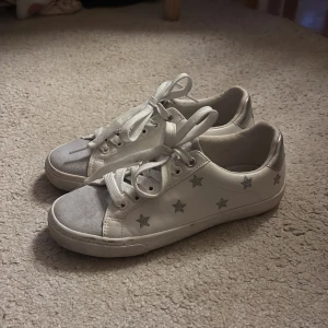Vita sneakers med silverstjärnor från Next - Snygga vita sneakers från Next med silvriga detaljer och broderade stjärnor på sidorna. Skorna har snörning, grå mockadetalj på tån och metallic-silver på hälen. Perfekta för dig som gillar en clean look med lite extra bling.