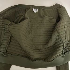 Olivgrön quiltad jacka från Burton London - Snygg olivgrön quiltad jacka från Burton London med raka sömmar och ribbade muddar. Jackan har dragkedja och praktiska fickor med zip. Perfekt för dig som gillar stilren och clean look.