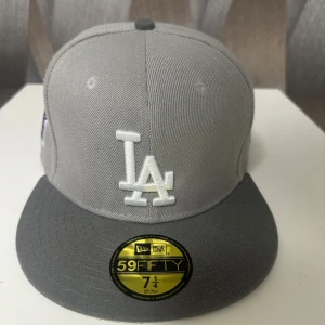 Grå LA Keps fitted - Snygg grå 59FIFTY keps från New Era med LA-logga broderad framtill i vitt. Kepsen har rak skärm, broderad detalj på sidan och klassisk 59FIFTY-stil. Tillverkad i polyester och bomull, perfekt för dig som gillar streetwear och sportig look.