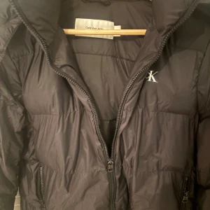 Svart pufferjacka från Calvin Klein Jeans - Svart pufferjacka från Calvin Klein Jeans med broderad CK-logga på bröstet. Jackan har hög krage, avtagbar huva och dragkedja framtill samt vid fickorna. Perfekt för kalla dagar och har en clean, stilren look.