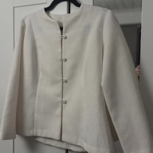 Beige kavaj med knappar - Stilren vit kavaj med rund halsringning och klädda knappar framtill. Jackan har en rak passform och är tillverkad i ett vävt tyg med diskret struktur. Perfekt för dig som gillar en clean och klassisk look. Figur sydd och hemmagjord🥰