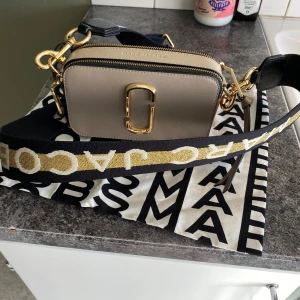 The snapshot, Marc Jacobs, beige med detaljer i guld och svart  - Snygg beige axelväska från Marc Jacobs med guldfärgade detaljer och dubbla dragkedjor. Väskan har en bred svart axelrem med guldtext och ett stilrent, rektangulärt format. Materialet är skinn och loggan syns tydligt framtill. Har inget slitage alls, och är oanvänd (fick den i present men har redan min favoritväska). Medföljer certifikat samt dustbag. 