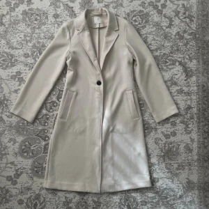 Beige kappa från H&M XS - Stilren beige kappa från H&M i storlek XS. Lång modell med klassisk krage, en knapp framtill och två fickor. Snyggt figursydd och är perfekt för dig som gillar en clean och elegant look. Perfekt höstkappa! Ing