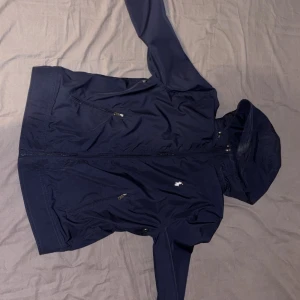 Mörkblå vindjacka från Polo Ralph Lauren - Snygg mörkblå vindjacka från Polo Ralph Lauren med huva, dragkedja och flera praktiska fickor med dragkedja. Jackan har det klassiska broderade Polo-loggan på bröstet och är tillverkad i lätt polyester. Perfekt för blåsiga dagar. Lägg gärna rimliga prisförslag