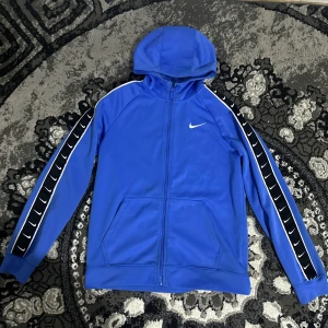 Blå Nike hoodie med dragkedja - Blå hoodie från Nike med hel dragkedja och huva. Svarta detaljer med vita Nike-loggor längs ärmarna och vita ränder. Två fickor framtill och klassisk sportig look. Perfekt för dig som gillar streetwear eller sportig stil. Storlek 147-158cm