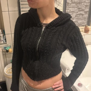 Svart croppad stickad hoodie med dragkedja - Svart croppad stickad hoodie med grov kabelstickning och dragkedja framtill. Tröjan har långa ärmar, bred ribbad mudd och en stor huva. Perfekt för dig som gillar en chill och trendig look under höst och vinter. Noppor finns men inget som stör😁