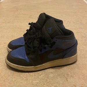 Nike Air Jordan 1 Mid svart/blå - Säljer ett par Nike Air Jordan 1 Mid i svart och blått skinn med klassisk swoosh och snörning. Skorna har en vit sula och ikoniska Jordan-loggor på sidan och plösen. Perfekta för dig som gillar streetstyle och vill sticka ut med en tidlös sneaker.