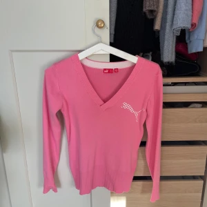 Rosa v-ringad tröja från Puma - Säljer en rosa v-ringad tröja från Puma med långa ärmar och ribbade muddar. Tröjan har en vit Puma-logga i prickigt mönster på bröstet. Perfekt för dig som gillar sportiga och stilrena plagg.