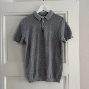 Grå pikétröja från Celio - Säljer en stilren grå pikétröja från Celio med klassisk krage och tre knappar framtill. Tröjan har korta ärmar och är tillverkad i ett mjukt, stickat material som känns skönt mot huden. Perfekt för dig som gillar en clean och enkel look.