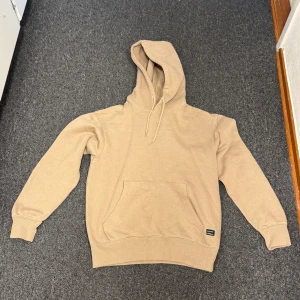 Beige jack&jones hoodie - Säljer en stilren beige hoodie med huva och snörning. Tröjan har en stor magficka framtill och ribbade muddar vid ärmslut och nederkant. Perfekt för en chill och avslappnad look.