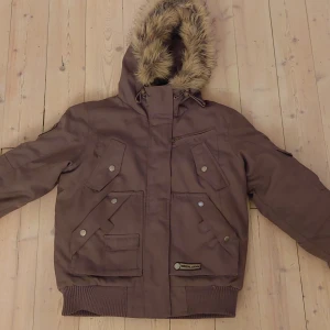 Brun vinterjacka med fuskpäls från Vero Moda - Snygg brun vinterjacka från Vero Moda med huva och fuskpäls. Jackan har flera fickor med tryckknappar, ribbade muddar och dragkedja framtill. Perfekt för kalla dagar med sin varma och rejäla känsla. Kommer från ett djur och rökfritt hem. 