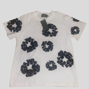 Denim Tears T-Shirt - Vit t-shirt från Denim Tears med unikt svart blommönster över hela plagget. Klassisk rund hals och korta ärmar. Tillverkad i mjuk bomull för skön känsla och snygg passform. Perfekt statement-piece för dig som vill sticka ut.