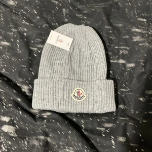 Grå mössa - Snygg grå ribbstickad mössa från Moncler med klassisk uppvikt kant och broderad logotyp framtill. Perfekt accessoar för kalla dagar och ger en clean look till din outfit. Tillverkad i mjukt material som håller dig varm.