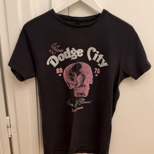 Mörkgrå t-shirt Jack & Jones med tryck - Mörkgrå t-shirt från Jack & Jones med coolt tryck framtill i vitt och rosa, med texten 'Dodge City' och en stor dödskalle. Klassisk passform med rund hals och korta ärmar. Tillverkad i mjuk bomull som är skön att bära. Storlek M.