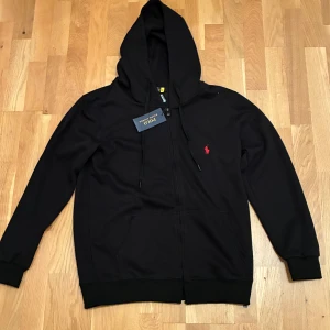 Svart hoodie från Polo Ralph Lauren - Svart hoodie från Polo Ralph Lauren med dragkedja, huva med snörning och rött broderat logomärke på bröstet. Tillverkad i mjuk bomullsmix, perfekt för chill dagar. Klassisk passform och ribbade muddar vid ärmslut och nederkant. Storlek XL för Barn men passar S för Vuxen