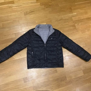 Svart och grå dunjacka från Polo Ralph Lauren - Snygg och lätt dunjacka från Polo Ralph Lauren som är vändbar med svart på ena sidan och grå på andra. Jackan har dragkedja framtill, två sidofickor med dragkedja och diskret logga på bröstet. Perfekt för kyliga dagar och enkel att matcha. Pris kan diskuteras