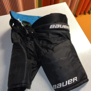 Svarta hockeyshorts Bauer JR Large - Säljer ett par svarta hockeyshorts från Bauer i juniorstorlek Large. Shortsen har snörning i midjan och vadderad insida med blått och grönt mönster. Bauer-logga på benet och midjan. Perfekta för hockeyträning eller match.