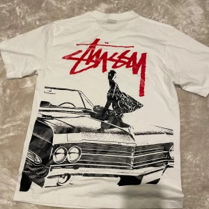 Vit Stüssy t-shirt - Vit t-shirt från Stüssy i 100% bomull. Stor grafisk print på ryggen med bil och person, samt röd Stüssy-logga både fram och bak. Klassisk passform och korta ärmar, perfekt för dig som gillar streetwear och snygga prints.