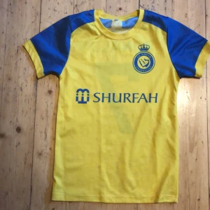 Al Nassr Ronaldo 7 fotbollströja gul/blå - Gul och blå fotbollströja från Al Nassr med Ronaldo 7 på ryggen. Tröjan har klubbmärke och sponsortryck på bröstet samt blå detaljer på axlar och ärmar. Tillverkad i lätt och bekvämt material, perfekt för fotbollsplanen eller att bära som supporter.