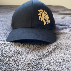 Svart keps med lejongult lejontryck - Det är den först släppta lion bar kepsen.       Det är limeted edition släppt från lion bars huvud kontor. Snygg svart keps med mesh på sidorna och justerbar snapback. Framtill finns ett stilrent lejongult lejontryck som ger kepsen en cool vibe. Perfekt accessoar för att lyfta din streetstyle. Materialet är en mix av bomull och syntet för bästa komfort. Det är den först släppta lion bar kepsen. 