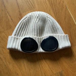 Vit ribbstickad co company mössa - Säljer en vit ribbstickad cp company mössa med svarta goggles framtill. Mössan har uppvikt kant och cool streetstyle-vibe, perfekt för dig som vill sticka ut. Materialet är mjukt och stretchigt, troligen bomull eller syntet. Pris kan diskuteras