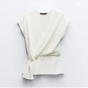 Vit Zara blus - Snygg vit omlottopp från Zara med knyt i sidan. Toppen har en elegant och enkel design med korta ärmar och mjukt fallande tyg. Perfekt att styla med jeans eller kjol för en fräsch look.