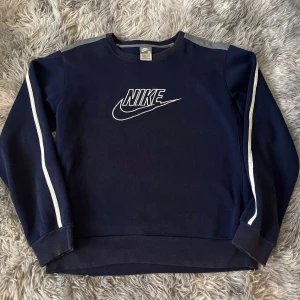Mörkblå Nike sweatshirt med vita detaljer - Snygg mörkblå sweatshirt från Nike med klassisk logga framtill. Tröjan har vita ränder längs ärmarna och grå axelpartier. Ribbstickade muddar vid ärmslut och nederkant. Perfekt för en sportig och avslappnad stil.