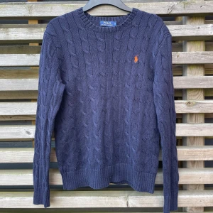 Mörkblå stickad tröja Polo Ralph Lauren - Kabelstickad mörkblå tröja från Polo Ralph Lauren med orange broderad logga på bröstet. Tröjan har rund halsringning, ribbade muddar och är långärmad. Perfekt för lager-på-lager under höst och vinter.