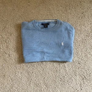 Ljusblå tröja från Ralph Lauren - Snygg ljusblå tröja från Polo Ralph Lauren i mjuk pima bomull. Klassisk rund halsringning, ribbade muddar och ikoniska broderade loggan på bröstet. Perfekt för dig som gillar stilrena och tidlösa plagg.