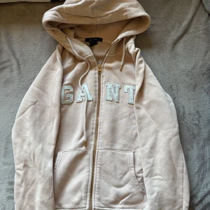 Beige hoodie från GANT med dragkedja - Snygg beige hoodie från GANT med broderad logga framtill. Tröjan har huva med snörning, dragkedja och två fickor framtill. Perfekt för en chill och avslappnad stil.