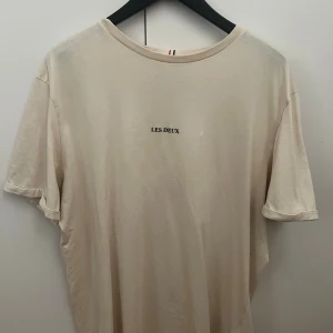 Beige t-shirt från Les Deux - Snygg beige t-shirt från Les Deux med minimalistisk svart text på bröstet. Klassisk rund halsringning och korta ärmar. Tillverkad i mjuk bomull som känns skön mot huden. Perfekt för en clean och stilren look.