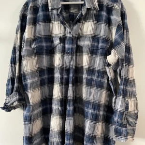 Blåvit rutig skjorta från H&M - Snygg, lång, rutig skjorta från H&M i blått och vitt med klassisk krage och knäppning framtill. Skjortan har två bröstfickor och långa ärmar som kan rullas upp. Perfekt för en avslappnad och cool stil.