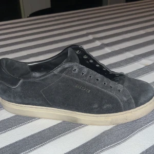 Svarta sneakers från Axel Arigato - Säljer ett par svarta sneakers från Axel Arigato i mocka med vit sula och diskret logga på sidan. Skorna har klassisk låg profil, rund tå och är utan snören. Insidan är fodrad med svart skinn för extra komfort. Inga skosnören.