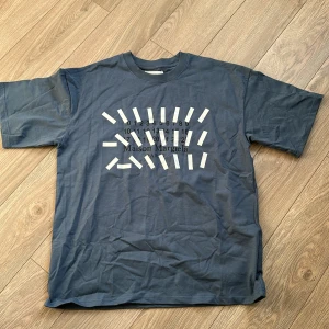 Blå Maison Margiela t-shirt med print i storlek L - Maison Margiela t-shirt i en snygg blå nyans med ikoniskt grafiskt print och logga framtill. Klassisk passform med rund hals och korta ärmar. Tillverkad i mjuk bomull och har diskreta vita stygn bak i nacken, typiskt för Margiela.