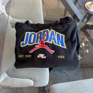 Jordan x Nike svart hoodie med tryck - Svart hoodie från Nike Jordan med stor broderad JORDAN-logga i blått och gult, röd Jumpman-figur och texten EST. 1985. Klassisk känguruficka, snörning i huvan och mjukt bomullsmaterial. Perfekt för dig som gillar streetwear och sportig stil. Det är en storlek S men känns som M