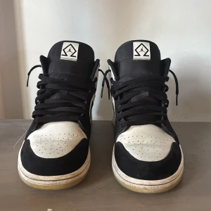 Nike Air Jordan 1 (Diamond Shorts) - Svartvita Nike Air Jordan 1 sneakers med klassisk design. Skorna har svart mocka och vitt skinn, svart swoosh och snörning. Ikonisk Air Jordan-logga på sidan och perforerad tå för extra stil. Perfekta för dig som gillar streetwear och vill sticka ut. (Original box medföljer)