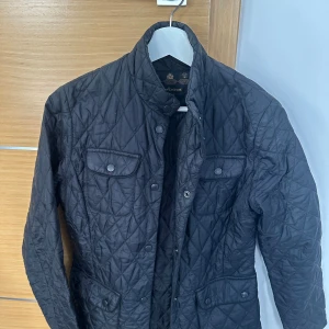 Svart quiltad jacka från Barbour - Quiltad svart jacka från Barbour med klassisk krage, tryckknappar och fickor framtill. Jackan har ett stilrent rutmönster och är tillverkad i polyester. Perfekt för dig som vill ha en tidlös och snygg look.