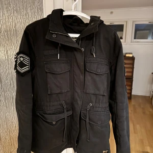 Svart parkajacka med patchar - Svart parkajacka från super dry med coola patchar på ärmarna och flera stora fickor framtill. Jackan har dragsko i midjan och huva, samt både dragkedja och knappar framtill. Passar dig perfekt nu inför hösten och om du har några funderingar är det bara att höra av sig🫡