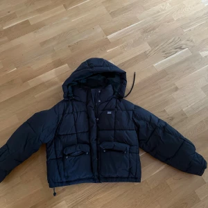 Svart pufferjacka från Levi's med huva - Säljer en svart pufferjacka från Levi's med huva och stora fickor framtill. Jackan har quiltad design, dragkedja och tryckknappar. Insidan är fodrad med mjuk fleece för extra värme. 