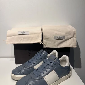 Valentino Garavani blå/vita sneakers - Snygga sneakers från Valentino Garavani i blått och vitt skinn. Klassisk låg modell med rund tå, snörning och ikoniska nitar på hälen. Skorna har en vit sula och stilren design som passar perfekt till streetwear eller casual outfits.