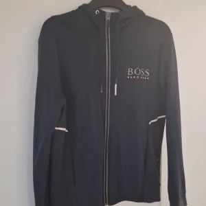 Svart hoodie från Hugo Boss med dragkedja - Svart hoodie från Hugo Boss med reflekterande detaljer och logga på bröstet. Tröjan har huva med snörning, hel dragkedja och fickor. Tillverkad i mjuk bomullsmix, perfekt för en chill och stilren look.