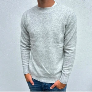 Ljusgrå stickad tröja herr - En stilren ljusgrå stickad tröja med rund hals och långa ärmar. Tröjan har en klassisk passform och ribbade muddar vid ärmslut och nederkant. Perfekt att matcha med jeans för en clean look.