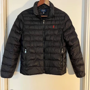 Svart pufferjacka från Polo Ralph Lauren - Svart pufferjacka från Polo Ralph Lauren med klassisk quiltad design och röd broderad logga på bröstet. Jackan har hög krage, dragkedja framtill och två fickor med dragkedja. Perfekt för kyliga dagar och riktigt snygg till streetwear.