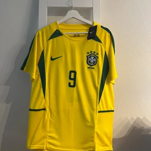 Brasilien Ronaldo 9 Nike fotbollströja M - Gul Brasilien fotbollströja från Nike med gröna detaljer, nummer 9 och Ronaldo på ryggen. Broderat landslagsemblem på bröstet och klassisk Nike-logga. Tillverkad i lätt och ventilerande polyester, perfekt för match eller samling.