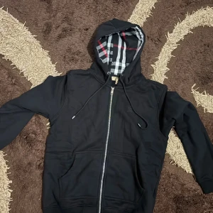 Svart hoodie från Burberry med dragkedja - Svart hoodie från Burberry med klassiskt rutigt foder i huvan. Tröjan har hel dragkedja, snörning i huvan och två fickor framtill. Stilen är clean och modern, perfekt för dig som gillar exklusiva streetwear-vibbar.