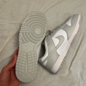 Nike Dunk vit/grå sneakers - Nike Dunk Low sneakers i en clean vit och ljusgrå färgkombination. Skorna har klassisk låg siluett, snörning och rund tå. Ovandelen är i slätt skinn med perforerade detaljer på tån och Nike-logga på hälen. Perfekta för dig som gillar stilrena och trendiga sneakers. Nytt skick bara testade inomhus! Köpta ifrån en annan från Vinted men passade ej längst fram lite för små där framme för mig.