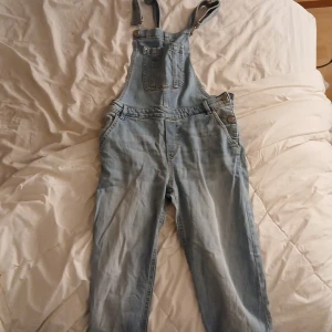 Ljusblå denim hängselbyxor Lindex - Snygga ljusblå denim hängselbyxor från Lindex med klassiska fickor fram och bak, justerbara axelband och raka ben. Perfekt för en avslappnad och trendig look. Tillverkade i bomull för skön känsla och stil.