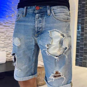 Rippade jeansshorts Jack & Jones - Säljer ett par ljusblå jeansshorts från Jack & Jones Originals med stora slitningar och rippade detaljer framtill. Klassisk femficksmodell, uppvikta benslut och normal passform. Perfekta för dig som gillar en avslappnad och trendig streetstil.