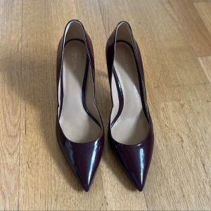Vinröda pumps från Michael Kors - Snygga vinröda pumps från Michael Kors i glansigt skinn. Skorna har spetsig tå och smal klack med en elegant gulddetalj längst ner. Perfekta för dig som vill ha en klassisk och stilren look med en touch av lyx.